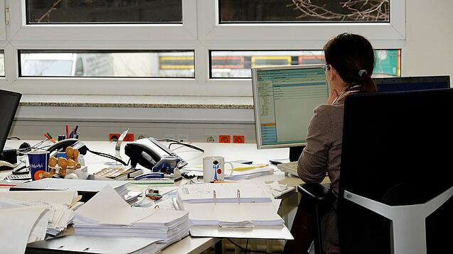 Eine Frau sitzt an einem Schreibtisch mit vielen Unterlagen und schaut auf einen Computer. 