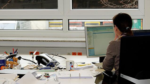 Eine Frau sitzt an einem Schreibtisch mit vielen Unterlagen und schaut auf einen Computer. 