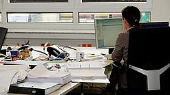 Eine Frau sitzt an einem Schreibtisch mit vielen Unterlagen und schaut auf einen Computer. 