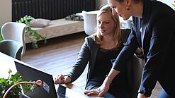 Zwei Frauen, eine sitzt, die andere stützt sich auf den Tisch, blicken auf einen Laptop, der auf dem Tisch vor den beiden steht.