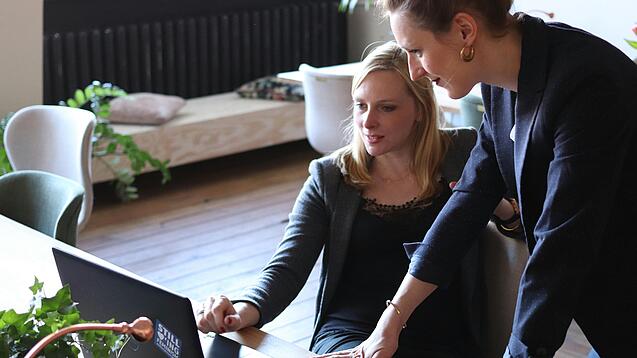 Zwei Frauen, eine sitzt, die andere stützt sich auf den Tisch, blicken auf einen Laptop, der auf dem Tisch vor den beiden steht.