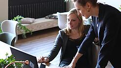 Zwei Frauen, eine sitzt, die andere stützt sich auf den Tisch, blicken auf einen Laptop, der auf dem Tisch vor den beiden steht.