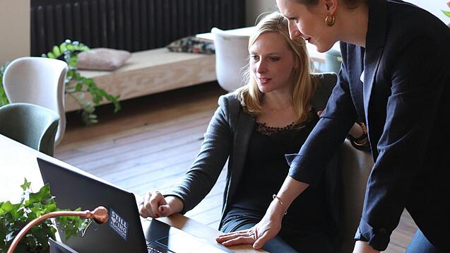 Zwei Frauen, eine sitzt, die andere stützt sich auf den Tisch, blicken auf einen Laptop, der auf dem Tisch vor den beiden steht.