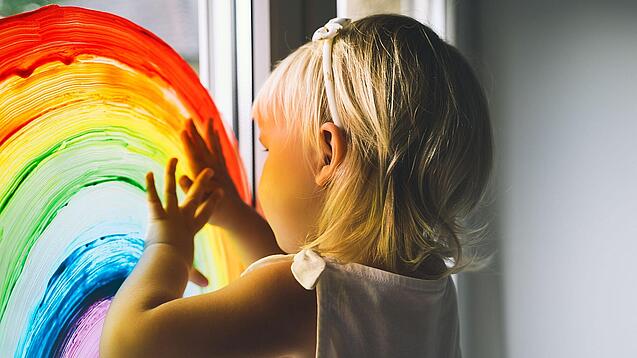 Blondes Kind malt mit Fensterfarben einen Regenbogen an das Fenster einer Kita.