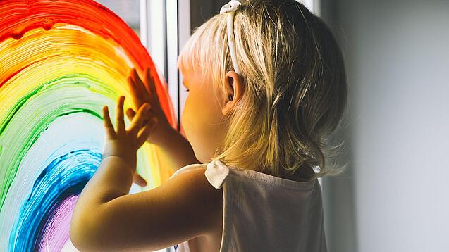 Blondes Kind malt mit Fensterfarben einen Regenbogen an das Fenster einer Kita.