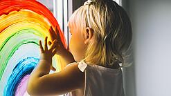 Blondes Kind malt mit Fensterfarben einen Regenbogen an das Fenster einer Kita.