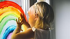 Blondes Kind malt mit Fensterfarben einen Regenbogen an das Fenster einer Kita.
