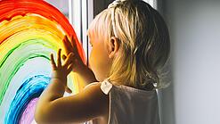 Blondes Kind malt mit Fensterfarben einen Regenbogen an das Fenster einer Kita.