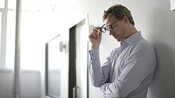 Ein Mann steht an eine Wand gelehnt und hält seine Brille in der Hand. Der Mann wirkt gestresst und erschöpft.