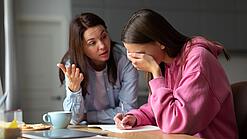 Eine junge Frau sitzt am Tisch und hält sich die Hand vor die Augen während eine andere Frau mit ihr redet.