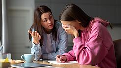 Jugendliche sitzt am Tisch und hält sich die Hand vor die Augen während eine Frau mit ihr redet.