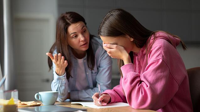 Jugendliche sitzt am Tisch und hält sich die Hand vor die Augen während eine Frau mit ihr redet.