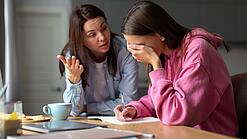 Jugendliche sitzt am Tisch und hält sich die Hand vor die Augen während eine Frau mit ihr redet.