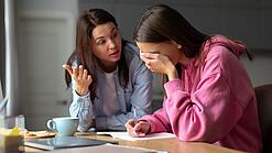 Jugendliche sitzt am Tisch und hält sich die Hand vor die Augen während eine Frau mit ihr redet.