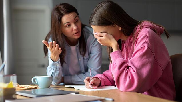 Jugendliche sitzt am Tisch und hält sich die Hand vor die Augen während eine Frau mit ihr redet.