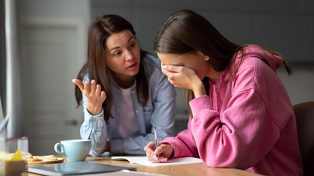 Jugendliche sitzt am Tisch und hält sich die Hand vor die Augen während eine Frau mit ihr redet.