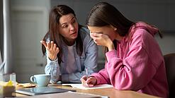 Jugendliche sitzt am Tisch und hält sich die Hand vor die Augen während eine Frau mit ihr redet.