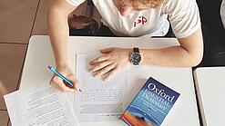 Ein Jugendlicher schreibt etwas auf einen Zettel, neben ihm liegt ein Englischwörterbuch.