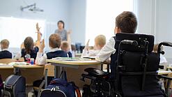 Es ist eine Schulklasse von hinten zu sehen, einer der Schüler sitzt in einem Rollstuhl. 