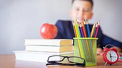 Tisch mit Schulbüchern, Stiften, Wecker und einem Jungen im Hintergrund