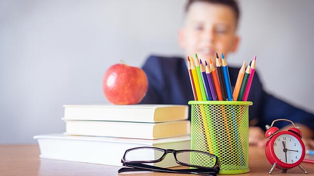 Tisch mit Schulbüchern, Stiften, Wecker und einem Jungen im Hintergrund