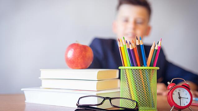 Tisch mit Schulbüchern, Stiften, Wecker und einem Jungen im Hintergrund