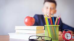 Tisch mit Schulbüchern, Stiften, Wecker und einem Jungen im Hintergrund