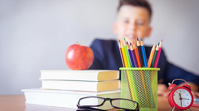 Tisch mit Schulbüchern, Stiften, Wecker und einem Jungen im Hintergrund