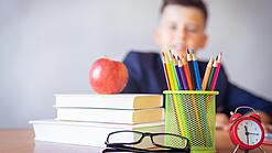 Tisch mit Schulbüchern, Stiften, Wecker und einem Jungen im Hintergrund