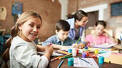 Ein Kind lächelt, während es mit anderen Kindern an einem Tisch sitzt und malt.