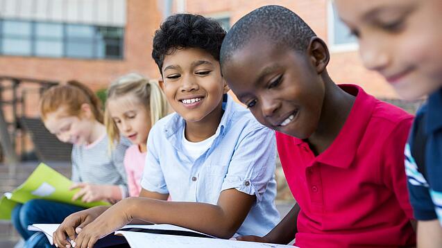 Mehrere Kinder schauen sitzend und lächelnd in Schulbücher und -hefte.
