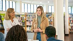 Eine junge Person und eine Lehrkraft stehen in einer Bibliothek und halten einen Vortrag.