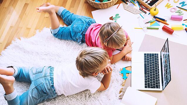 Kinder liegen auf einem Teppich und schauen auf einen Laptop.
