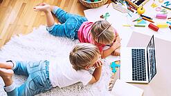 Kinder liegen auf einem Teppich und schauen auf einen Laptop.