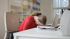 Frau sitzt am Schreibtisch und hat den Kopf auf die Tastatur ihres Laptops gelegt.
