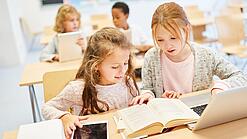 Zwei Mädchen sitzen in einem Klassenraum vor einem Laptop, einem Tablet und einem Buch
