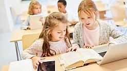 Zwei Mädchen sitzen in einem Klassenraum vor einem Laptop, einem Tablet und einem Buch