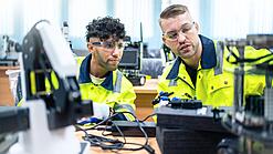 Zwei junge Männer mit Schutzbrillen und in gelben Warnjacken sitzen in einem Technik-Raum