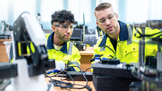 Zwei junge Männer mit Schutzbrillen und in gelben Warnjacken sitzen in einem Technik-Raum