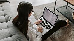 Eine Studentin sitzt auf dem Boden an ein Sofa angelehnt und arbeitet an einem Laptop, der auf ihrem Schoß liegt.