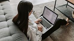 Eine Studentin sitzt auf dem Boden an ein Sofa angelehnt und arbeitet an einem Laptop, der auf ihrem Schoß liegt.