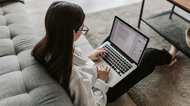 Eine Studentin sitzt auf dem Boden an ein Sofa angelehnt und arbeitet an einem Laptop, der auf ihrem Schoß liegt.