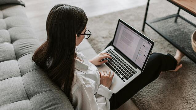 Eine Studentin sitzt auf dem Boden an ein Sofa angelehnt und arbeitet an einem Laptop, der auf ihrem Schoß liegt.