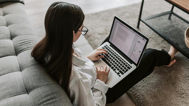 Eine Studentin sitzt auf dem Boden an ein Sofa angelehnt und arbeitet an einem Laptop, der auf ihrem Schoß liegt.