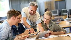Ein Lehrer zeigt einer Gruppe Schülerinnen und Schüler etwas auf einem Tablet.