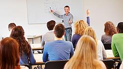 Lehrer steht vor der Klasse an einem Whiteboard und erklärt etwas