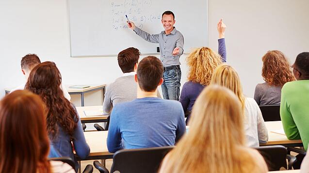Lehrer steht vor der Klasse an einem Whiteboard und erklärt etwas
