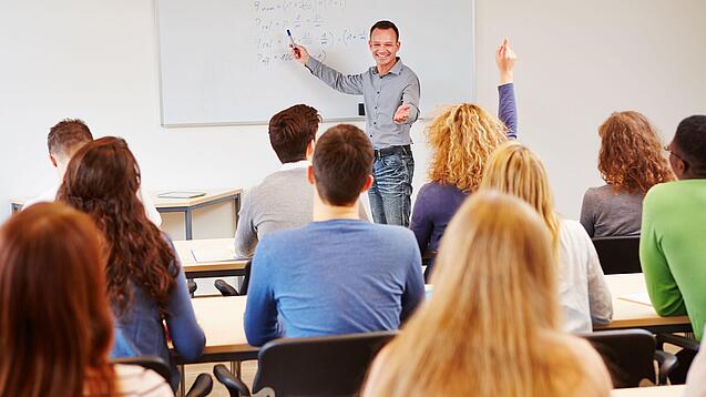 Lehrer steht vor der Klasse an einem Whiteboard und erklärt etwas