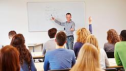 Lehrer steht vor der Klasse an einem Whiteboard und erklärt etwas