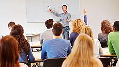 Lehrer steht vor der Klasse an einem Whiteboard und erklärt etwas